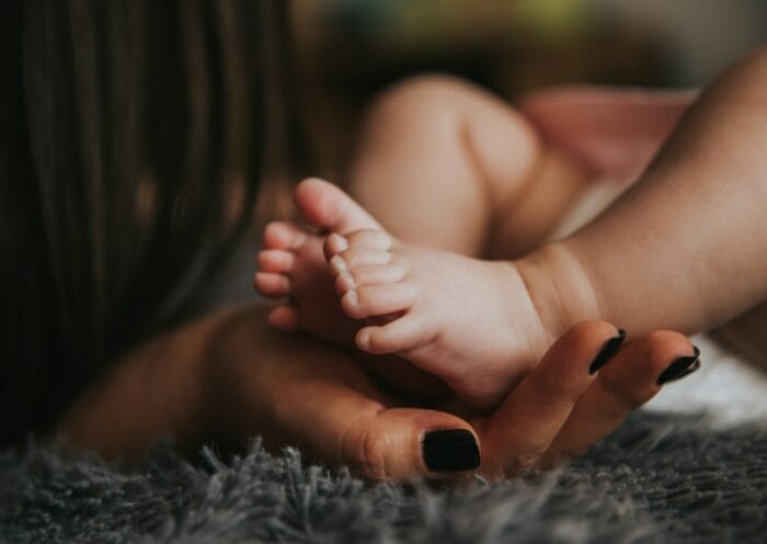 Infant's feet being held by a woman's hand with painted and manicured hands resting on a gray blanket