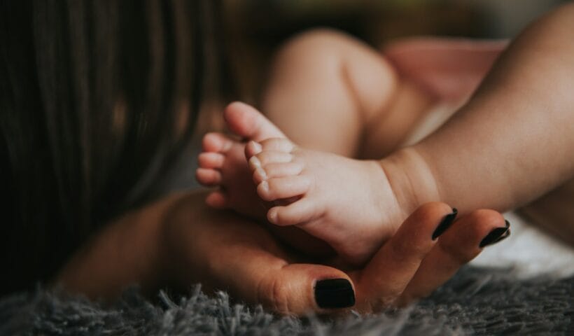 Infant's feet being held by a woman's hand with painted and manicured hands resting on a gray blanket